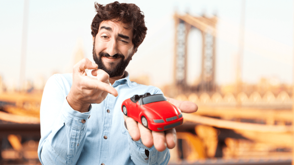 Man giving a creepy smile holding a toy car in one hand and pointing figure to the toy car with another hand.